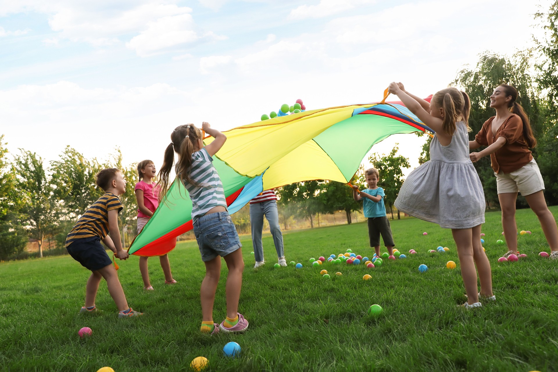 Group of children and teacher playing with rainbow playground parachute on green grass. Summer camp activity